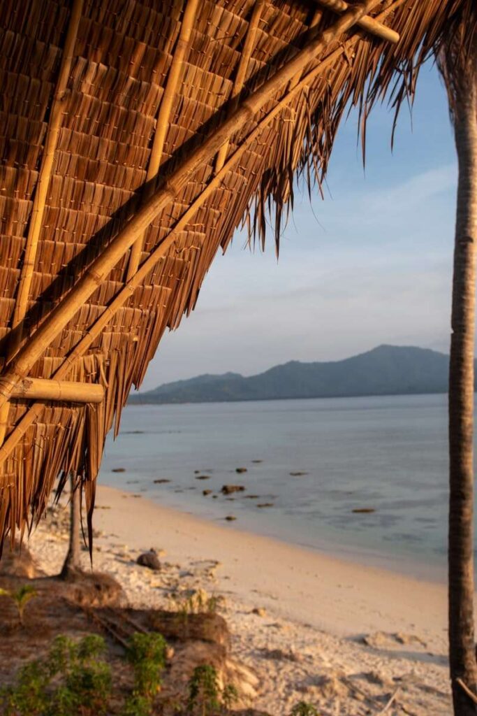 A thatched roof hut overlooks a tranquil sandy beach with scattered rocks. Embrace the Isla Experience as calm blue water stretches toward distant green mountains under a clear sky at sunset.