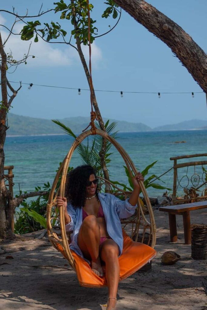 A woman with curly hair enjoys an Isla Experience, relaxing on a hanging wicker chair with an orange cushion at the beach. She wears sunglasses and a light blue shirt over a pink outfit, with the ocean and mountains in view.