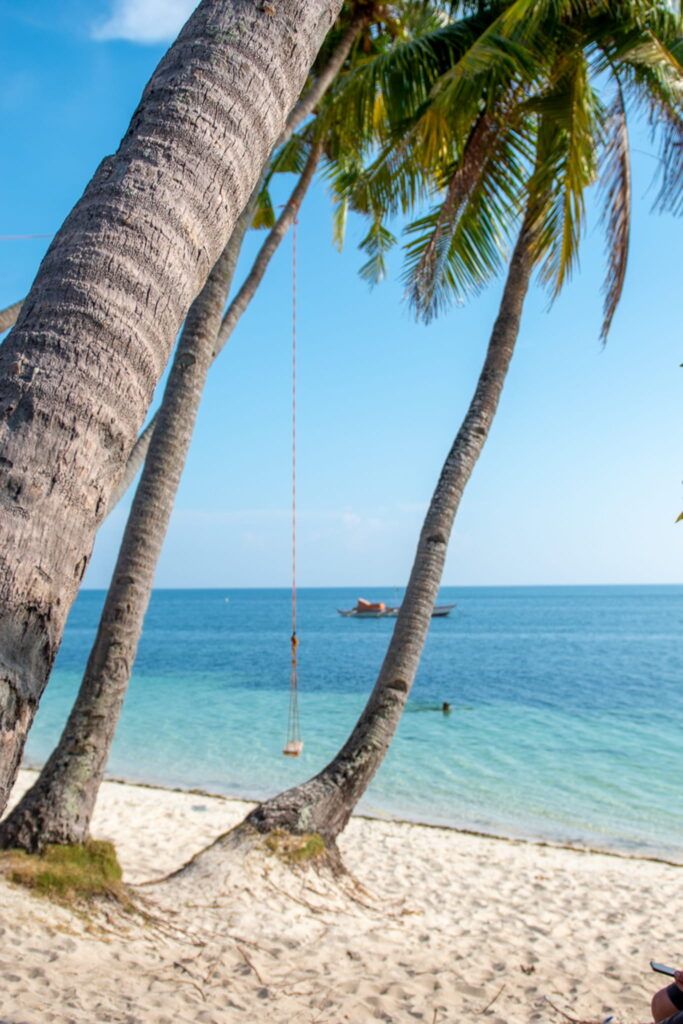 Sandy beach with leaning palm trees, a rope swing hanging from one tree, clear turquoise water, and a small boat floating on the calm sea under a bright blue sky—perfect for your Philippines island-hopping itinerary.