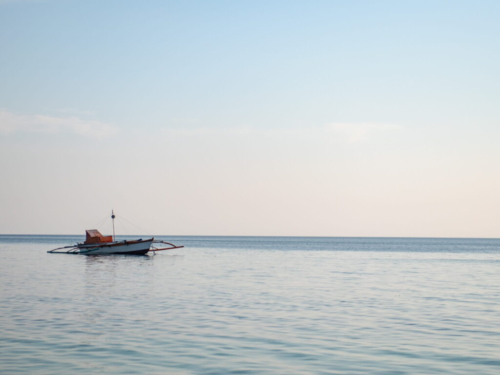 A small wooden boat floats on calm, clear water under a pale blue sky with soft clouds—perfect for a peaceful Philippines island-hopping itinerary. The horizon is visible, evoking a serene and tranquil atmosphere.