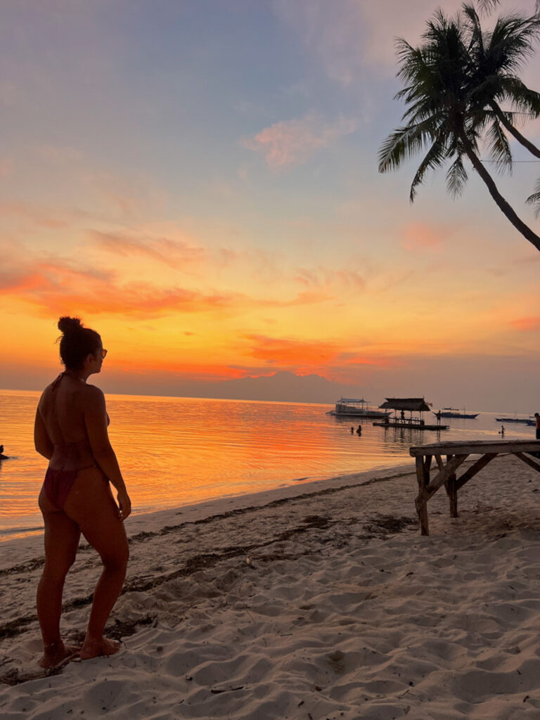 A woman in a bikini stands on a sandy beach at sunset, looking toward the colorful sky and calm sea—one of the stunning places to visit in Siquijor, where palm trees and wooden huts are silhouetted against the horizon.