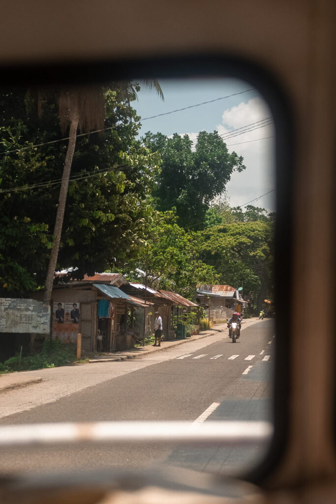 View through a vehicle window showing a rural road with a motorcyclist, roadside houses with metal roofs, lush green trees, and a partly cloudy sky—an inviting glimpse into daily life on a Philippines island-hopping itinerary.