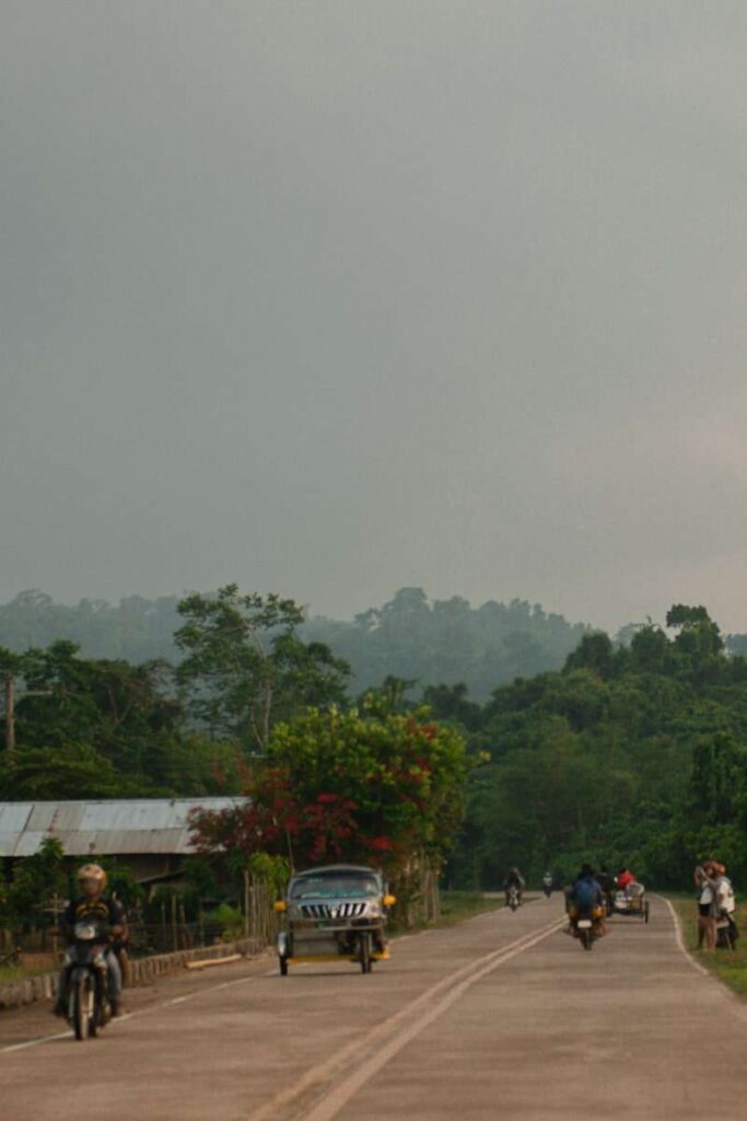 A rural road with several people riding motorcycles and a car driving, bordered by lush green trees and plants, set under a cloudy sky with hills in the background—perfect scenery for your Philippines island-hopping itinerary.