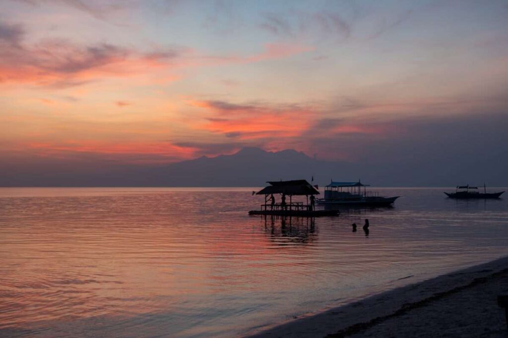 A tranquil sunset over calm sea waters, with vibrant orange and pink clouds. Silhouettes of boats suggest a Philippines island-hopping itinerary, while two people stand in the shallow water near a sandy shore and a small hut on stilts.