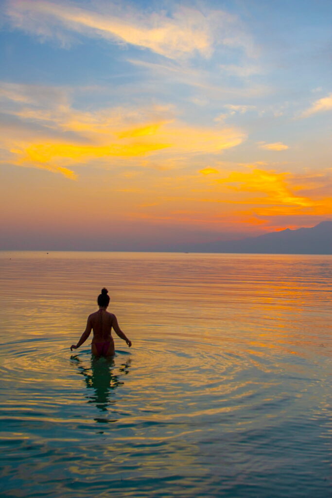 A person stands waist-deep in calm water at sunset, facing the horizon—an idyllic scene to end a Philippines island-hopping itinerary. The sky glows with orange, yellow, and blue hues, reflecting on the rippling surface of the water.