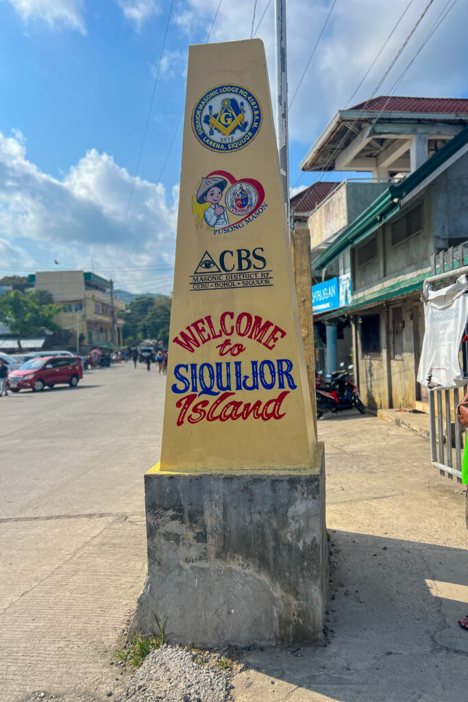 A tall concrete welcome marker on a street reads “Welcome to Siquijor Island,” greeting travelers who seek new places to visit in Siquijor. Various local organization logos, nearby buildings, cars, and a blue sky complete the scene.