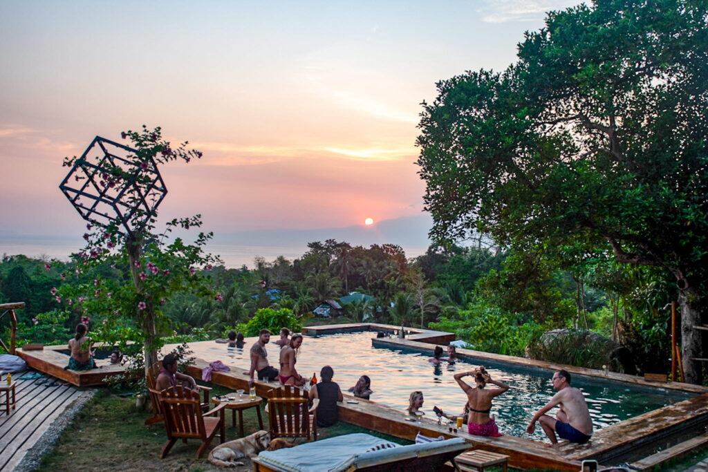 People relax and swim in an outdoor pool surrounded by trees and wooden furniture at sunset, the perfect end to a Philippines island-hopping itinerary, with the sun dipping behind distant hills and the ocean in the background.