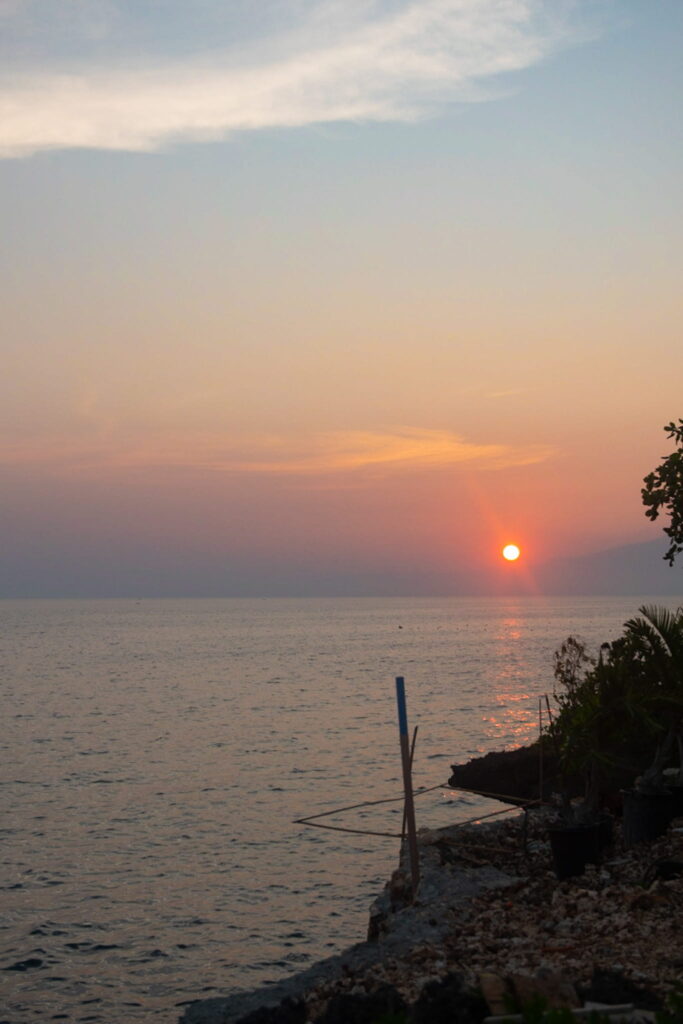 The sun sets over calm ocean waters, casting an orange glow on the horizon—a perfect scene for your Philippines island-hopping itinerary, with silhouetted plants and rocks lining the shore under a partly cloudy sky.