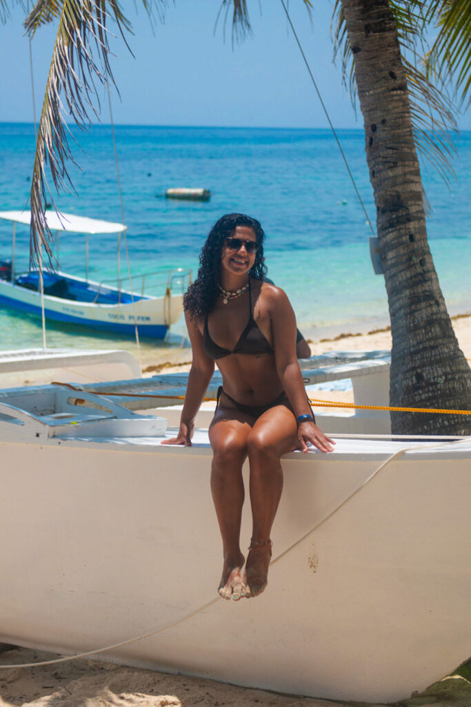 A woman wearing a black bikini sits on a white boat beached on sandy shore, surrounded by palm trees and clear turquoise water—one of the stunning places to visit in Siquijor, with another boat visible in the background on a sunny day.