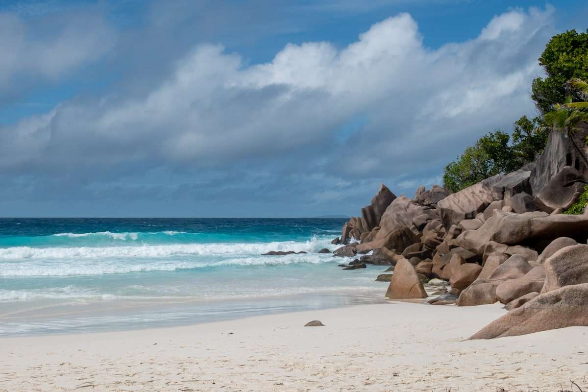 A serene beach scene featuring clear blue water gently lapping against a sandy shore, with unique, large rock formations and lush greenery in the background. The sky is partly cloudy, enhancing the tranquil atmosphere—an ideal start to any 5-day Seychelles itinerary.