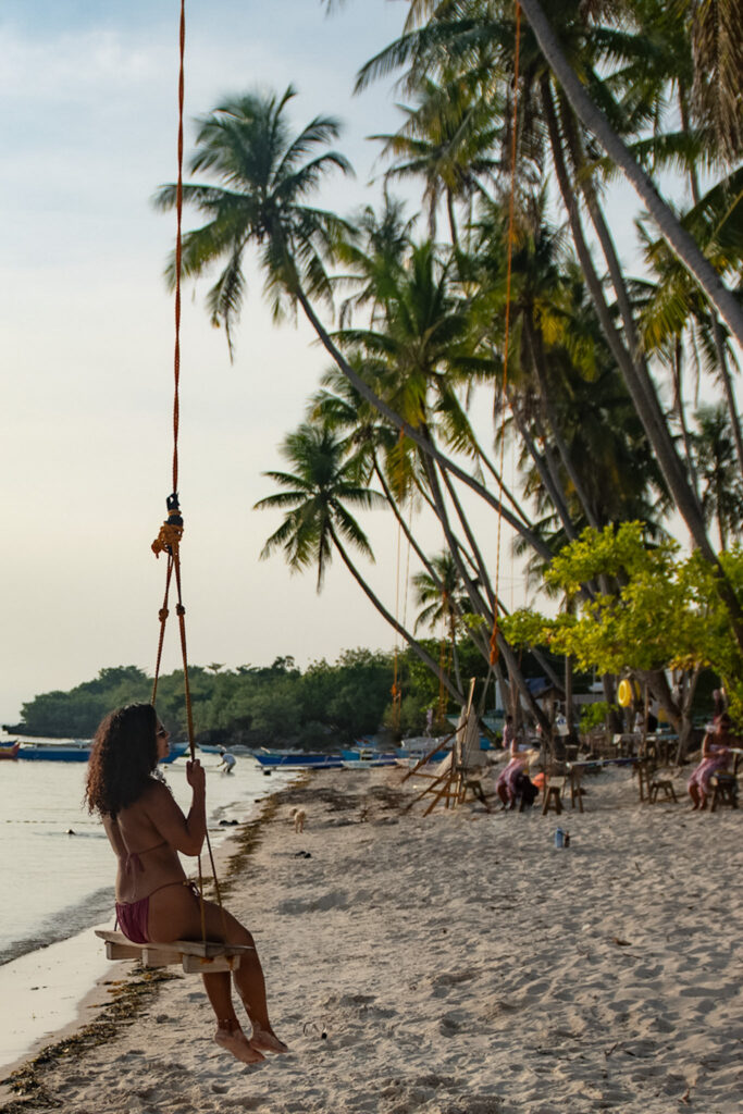 A woman in a swimsuit sits on a swing hanging from a palm tree on a sandy beach, one of the picturesque places to visit in Siquijor, with other people and swaying palms lining the shoreline in the background.