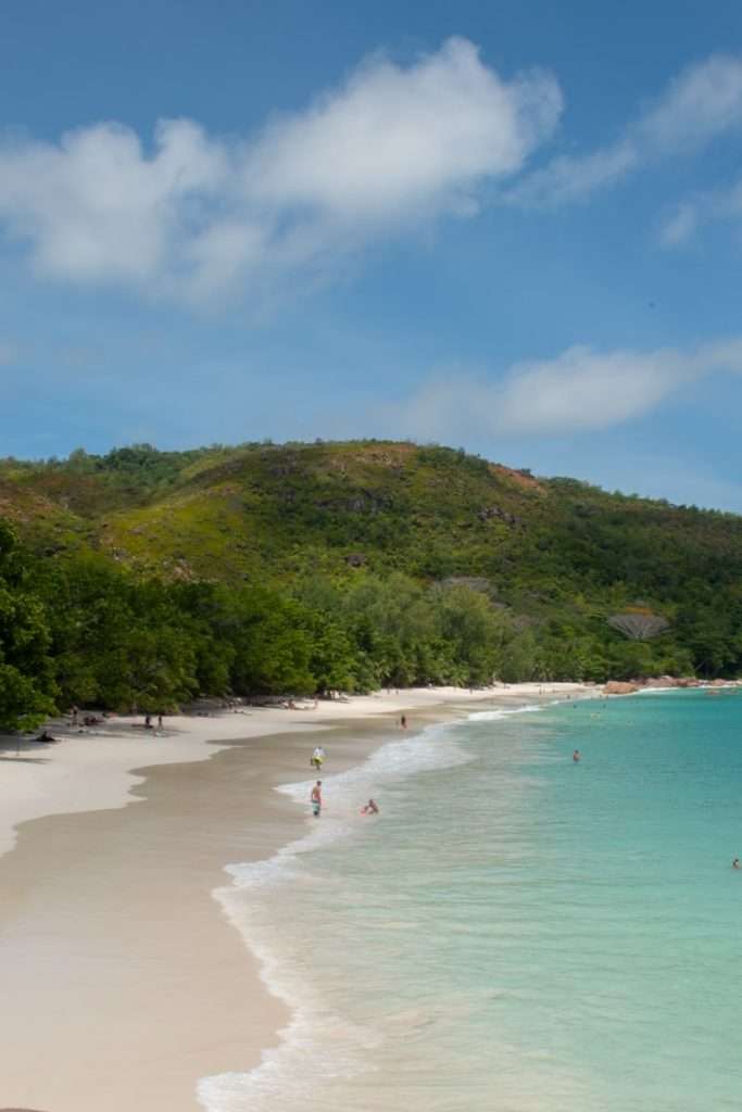 A serene beach scene with clear turquoise water gently lapping onto the white sandy shore. A few people are enjoying the water and the beach under a bright blue sky with scattered clouds. Lush green hills rise in the background, covered with dense vegetation. Welcome to Anse Lazio Beach, featured on every 5-day Seychelles itinerary.