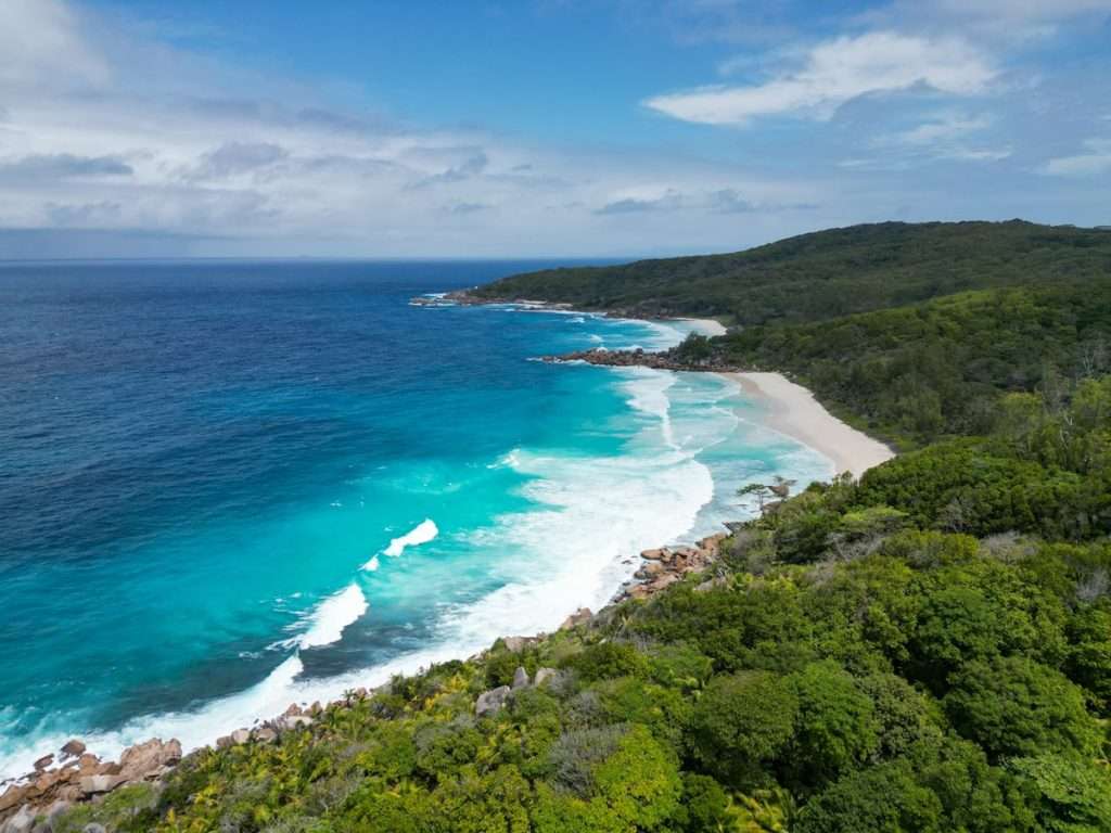 A breathtaking view of a pristine tropical beach with turquoise waters and white waves crashing onto the sandy shore. Lush green vegetation covers the surrounding hills, while the sky is partly cloudy, creating a serene and picturesque scene; perfect for your 5-day Seychelles itinerary including Anse Cocos on La Digue.