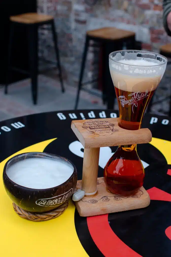 A glass of amber beer with a foamy head sits in a wooden holder next to a ceramic cup filled with frothy liquid on a colorful table with bold red, yellow, and black patterns. Two bar stools are in the background.