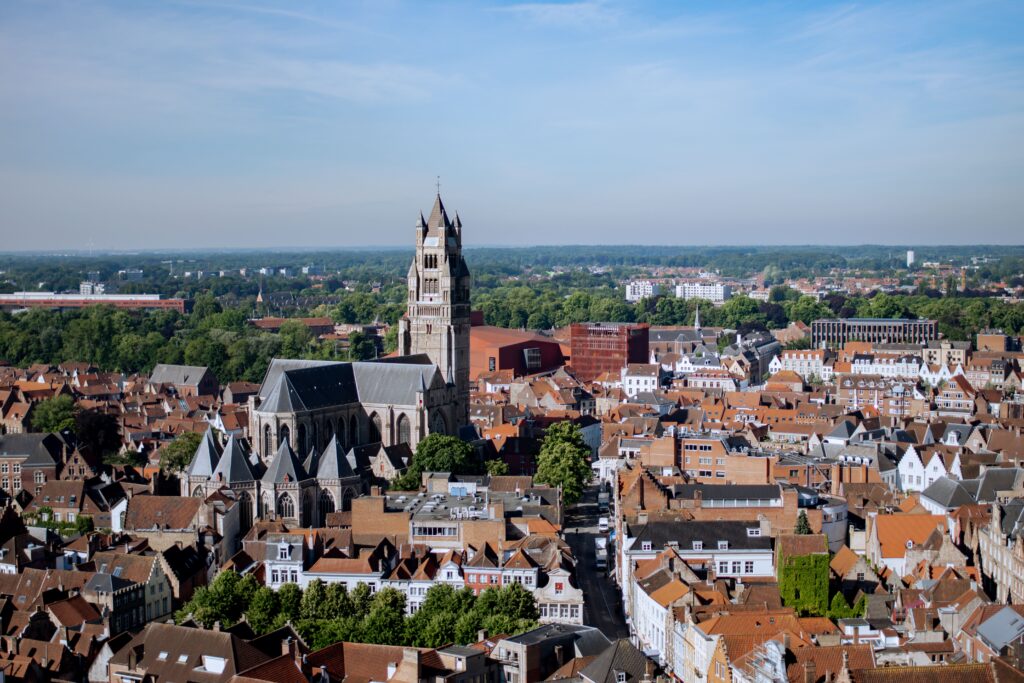 Aerial view of a European city with red-roofed buildings, a prominent gothic church with a tall tower in the center, and green trees in the background under a clear blue sky.