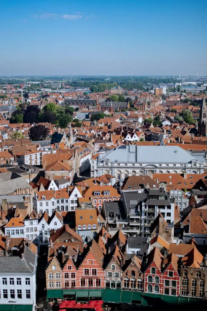A panoramic view of Bruges, Belgium, showing rows of colorful historic buildings with red-tiled roofs under a clear blue sky. In the foreground are stepped-gable houses lining a square.