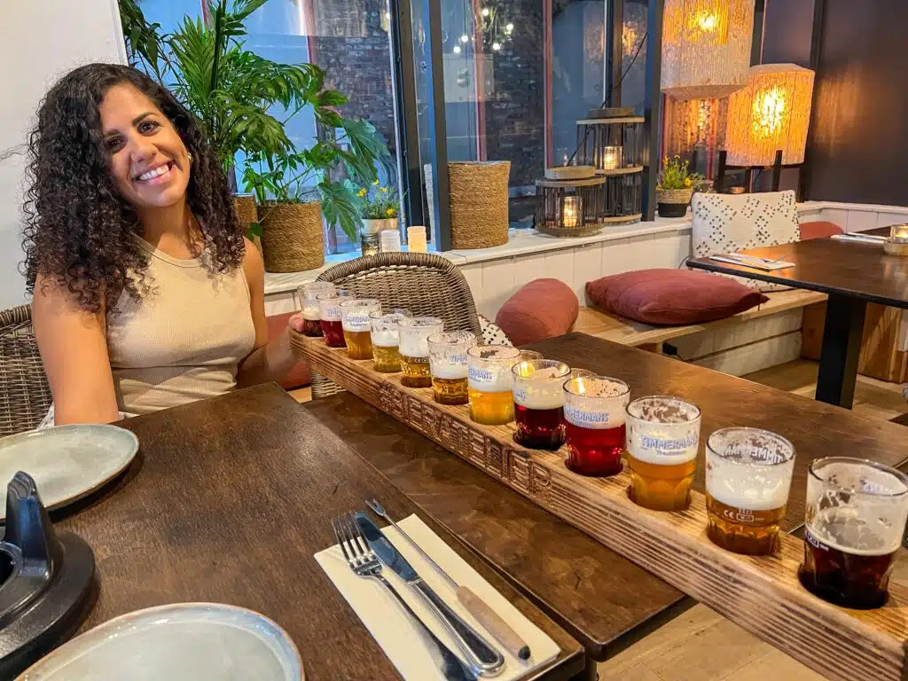 A woman with curly hair smiles at a table in a cozy restaurant, sitting beside a long wooden tray holding a colorful beer flight with multiple small glasses of different beers. The setting has plants and warm lighting.