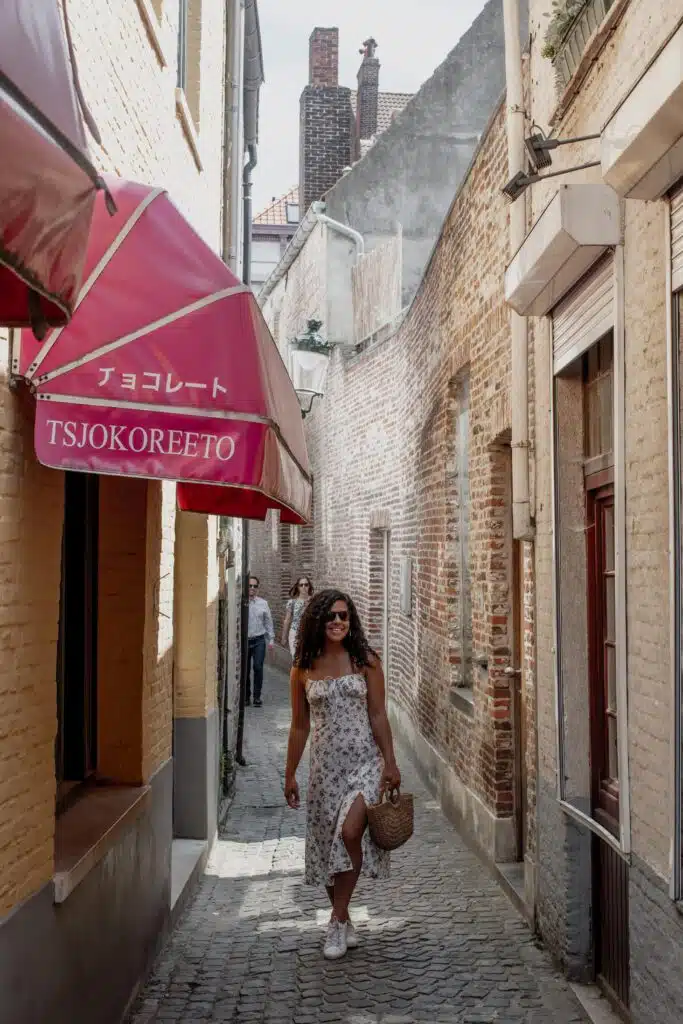 A woman in a white floral dress and sunglasses walks down a narrow cobblestone alley with brick walls, under a pink awning with Japanese and English text reading “TSJOKOREETO.”.