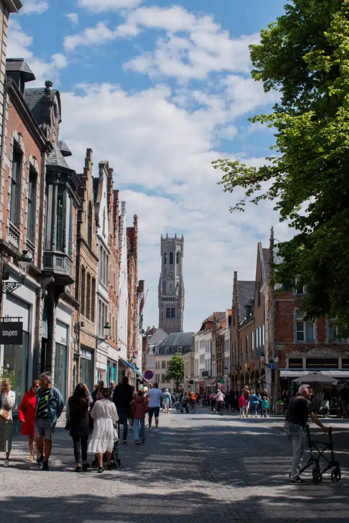 A cobblestone street in a European city with people walking and biking, lined with historic brick buildings, and a tall clock tower visible in the background under a partly cloudy sky.