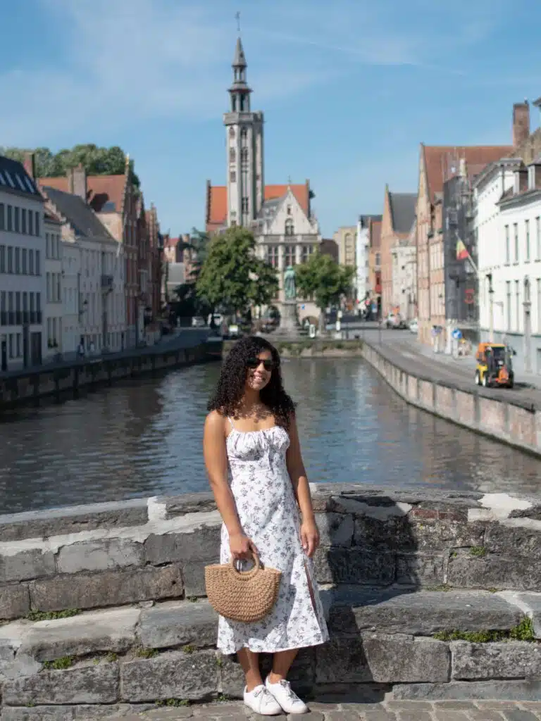 A woman in a white floral dress and sunglasses stands on a stone bridge over a canal in a European city, with historic buildings and a tall clock tower visible in the background under a clear blue sky.