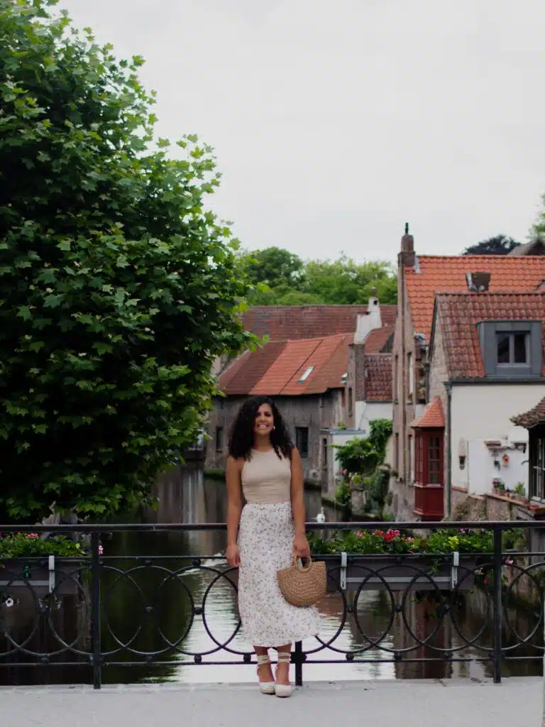 A woman in a light sleeveless top and floral skirt stands on a bridge in front of a canal, with quaint houses and a leafy tree in the background. She is holding a straw bag and smiling.