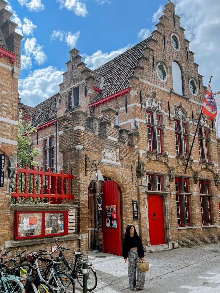 A woman walks past a historic brick building with red-trimmed windows and doors, a flag, and several parked bicycles on a cobblestone street under a partly cloudy sky.
