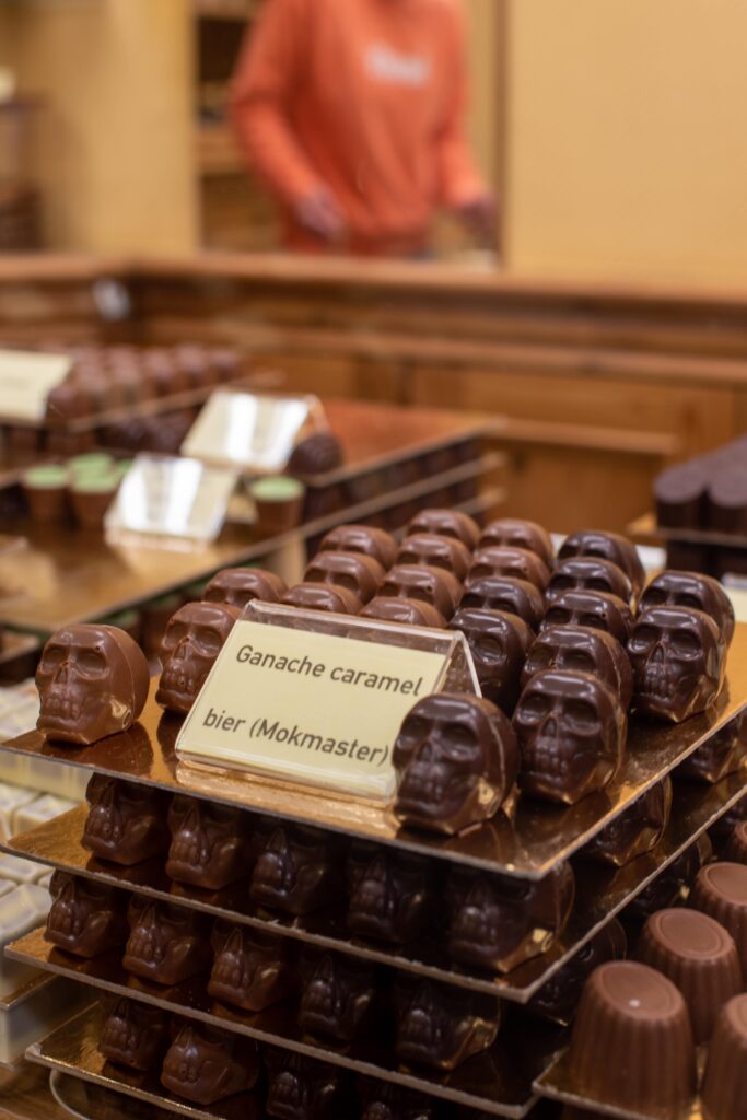 Rows of skull-shaped chocolates are displayed on trays in a shop, with a sign reading "Ganache caramel bier (Mokmaster)" in front. A person in an orange shirt is blurred in the background.
