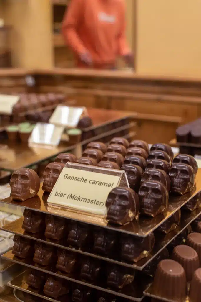 Rows of skull-shaped chocolates are displayed on trays in a shop, with a sign reading "Ganache caramel bier (Mokmaster)" in front. A person in an orange shirt is blurred in the background.