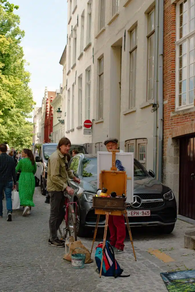 A street artist with a hat paints at an easel on a cobblestone street near parked cars, while another person stands beside a bicycle. People walk in the background, and tall buildings line the street.