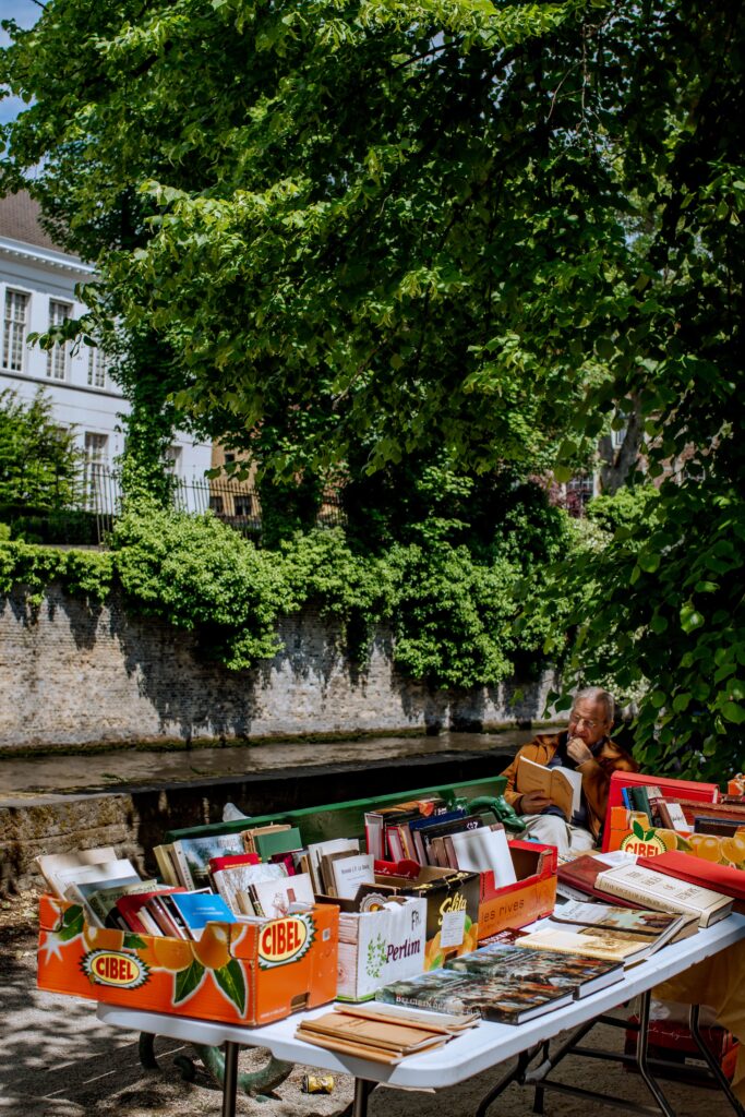 A man sits at an outdoor table under leafy trees, surrounded by boxes of books for sale. In the background, there's a canal and white buildings partially covered with greenery.