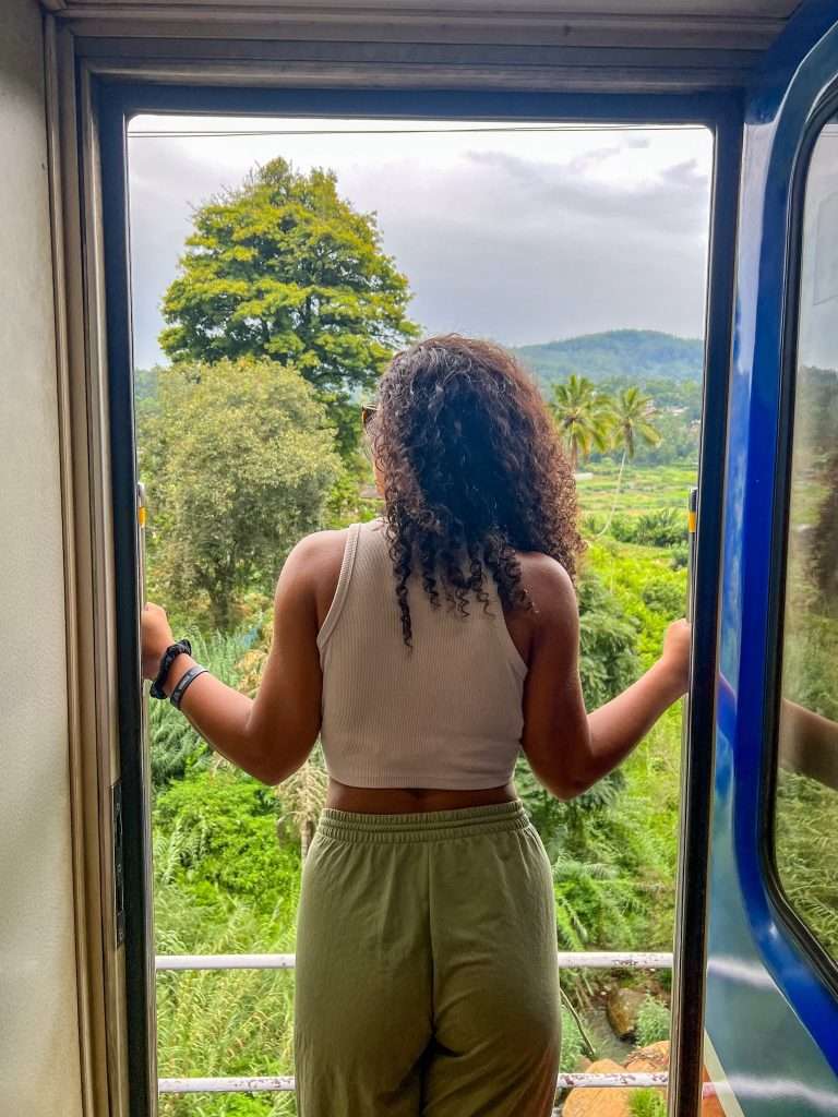 A person with curly hair, wearing a white tank top and green pants, stands at the open door of the train traveling from Colombo to Ahangama, looking out at a lush green landscape with trees, hills, and overcast skies.