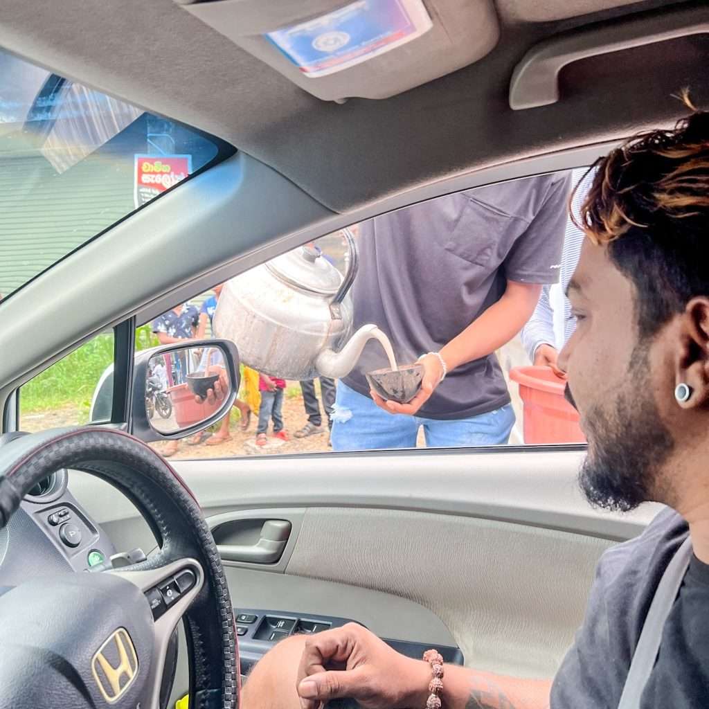 A man in a car on the bustling Colombo to Ahangama route receives tea from someone outside pouring from a metal teapot. The open window reveals the cozy interior, while the backdrop showcases a building and a lively group of people.