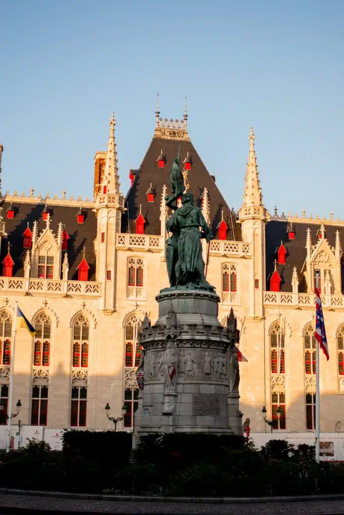 A bronze statue stands in front of a historic, ornate building with pointed towers, spires, and red window shutters, illuminated by warm sunlight. Flags are visible around the base of the statue.
