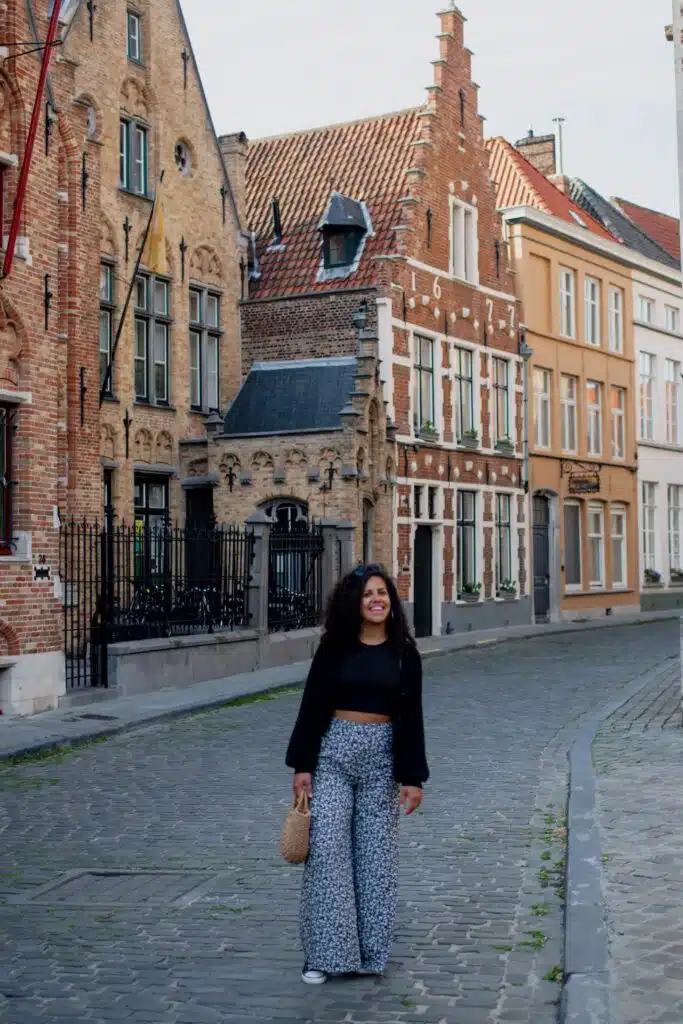 A woman with curly hair, wearing a black jacket and patterned pants, stands smiling on a cobblestone street lined with old, ornate brick buildings. The street is quiet and empty, with European architecture in the background.