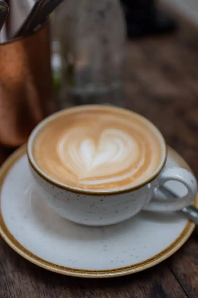 A close-up of a cup of latte with heart-shaped latte art on top, served in a white speckled ceramic cup and saucer on a wooden table. A spoon rests on the saucer and utensils are in the blurred background.