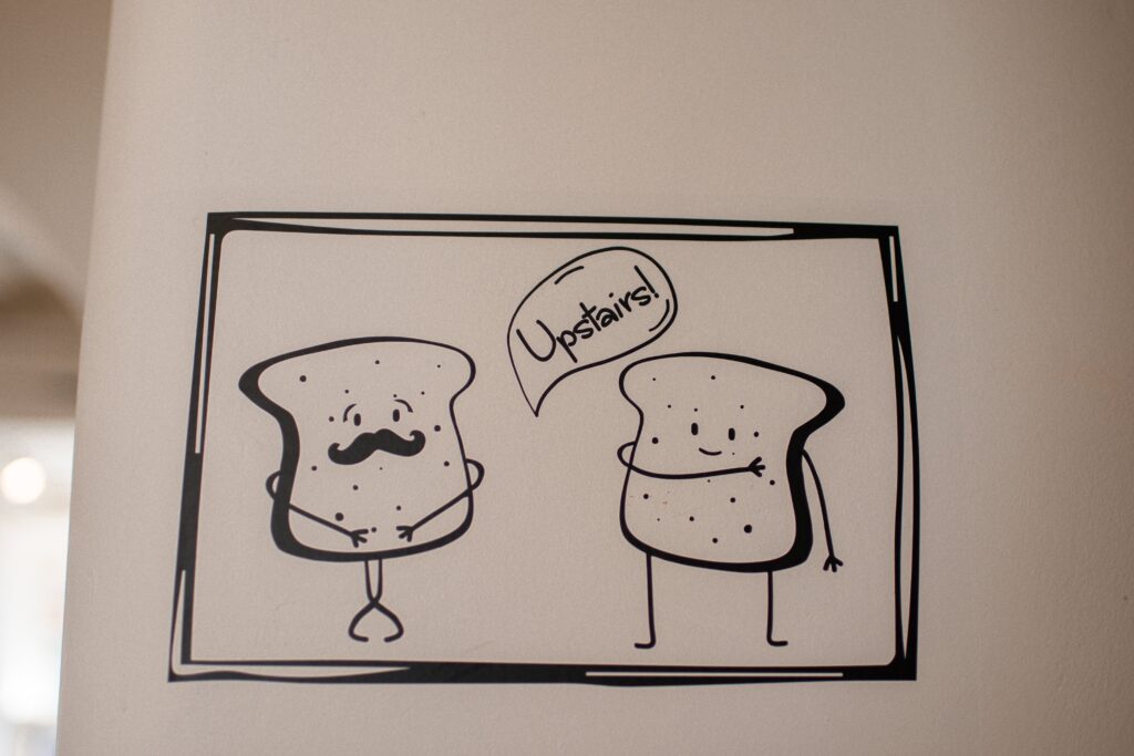 A black-and-white cartoon drawing of two slices of bread on a wall; one has a mustache and crossed legs, while the other points upward with a speech bubble that says "Upstairs!.