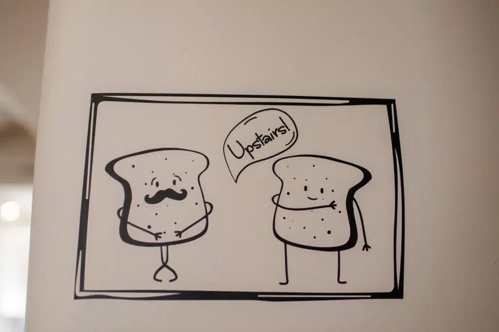 A black-and-white cartoon drawing of two slices of bread on a wall; one has a mustache and crossed legs, while the other points upward with a speech bubble that says "Upstairs!.