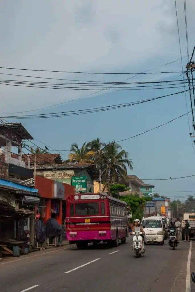 A bustling street scene captures a pink bus making its way from Colombo to Ahangama, weaving through motorcycles on a busy road flanked by vibrant shops and buildings. Overhead, tangled wires cross the cloudy sky while palm trees sway gently in the breeze.