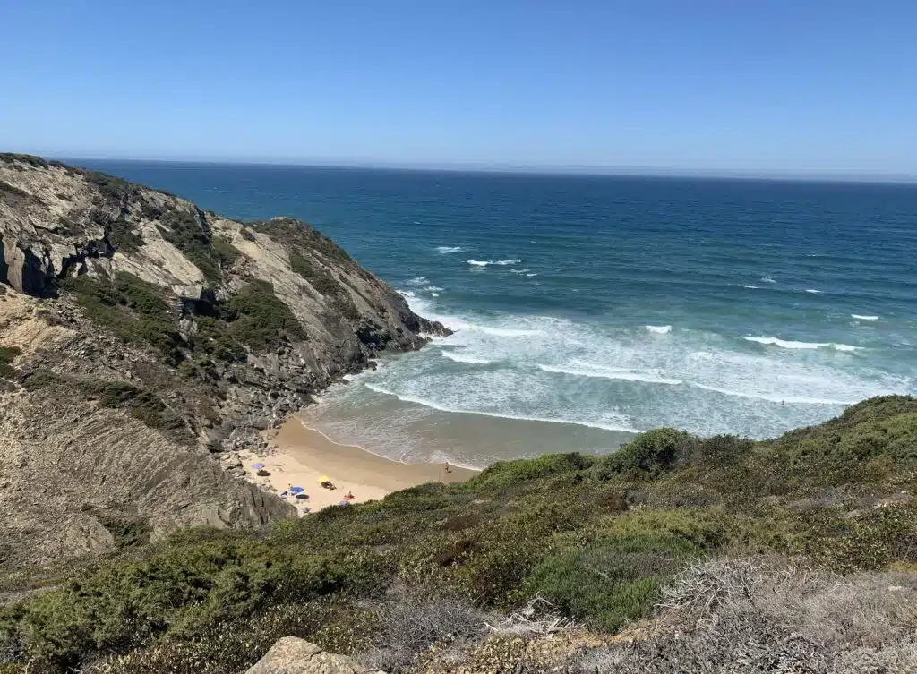 A secluded sandy beach with turquoise waves, bordered by rocky cliffs and green vegetation under a clear blue sky—one of the top things to do in Sagres. A few people and colorful umbrellas add life to the peaceful shore.