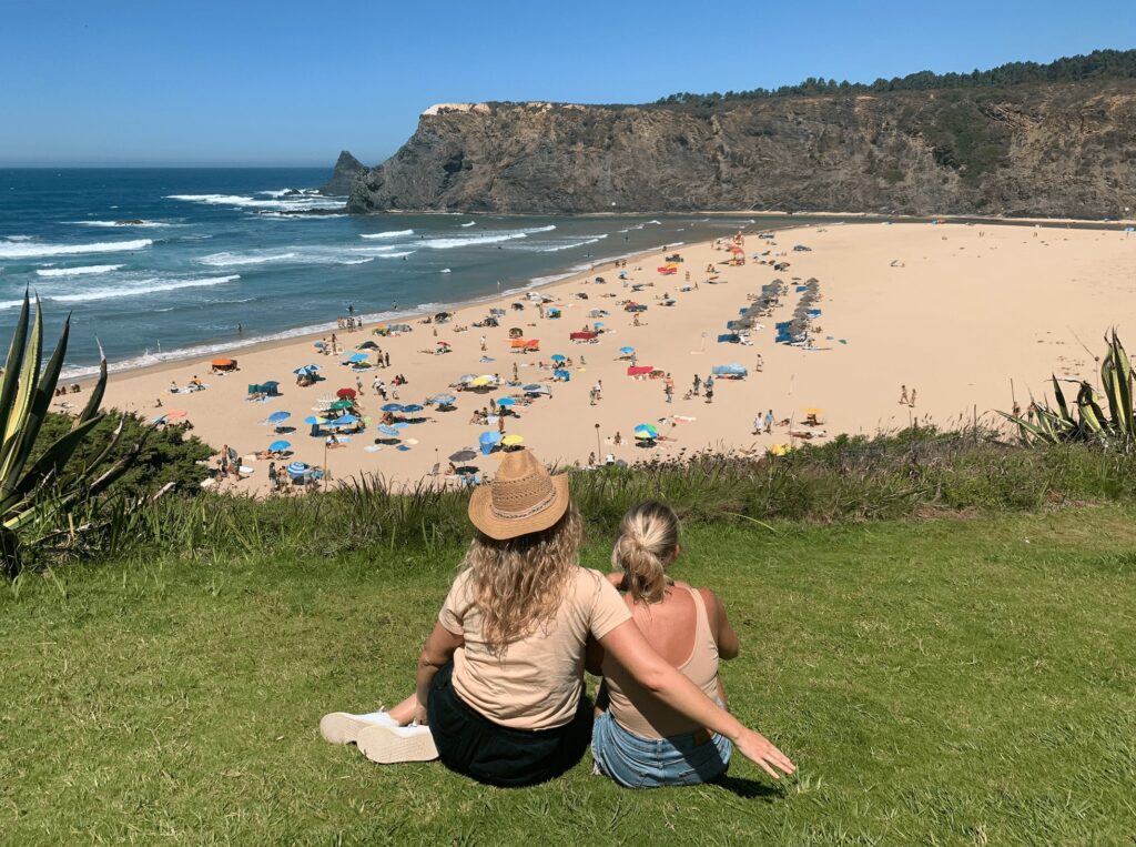 Two women sit on grass overlooking a sandy beach in Sagres, with colorful umbrellas and people below. Waves roll in from the blue ocean, rocky cliffs rise in the background—a perfect spot to enjoy some of the top things to do in Sagres under a clear sky.