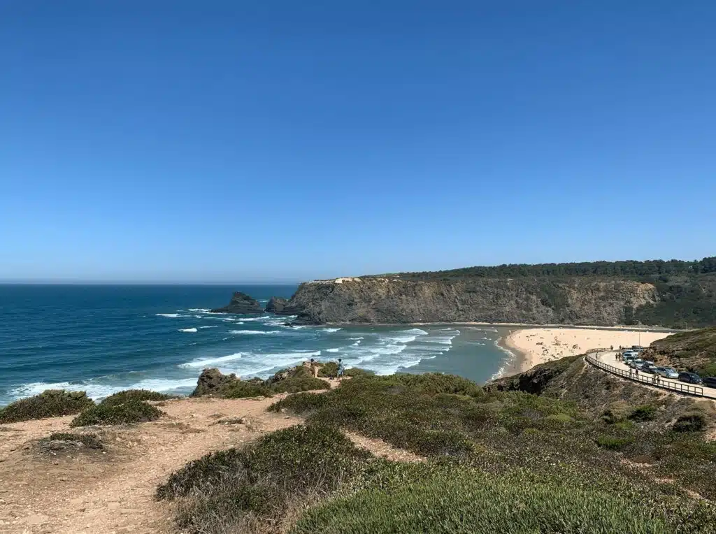 A scenic view of a sandy beach in Sagres bordered by cliffs and green vegetation, with waves crashing onto the shore under a clear blue sky—a perfect spot to explore among the many things to do in Sagres. A winding path and parked cars are visible on the right.