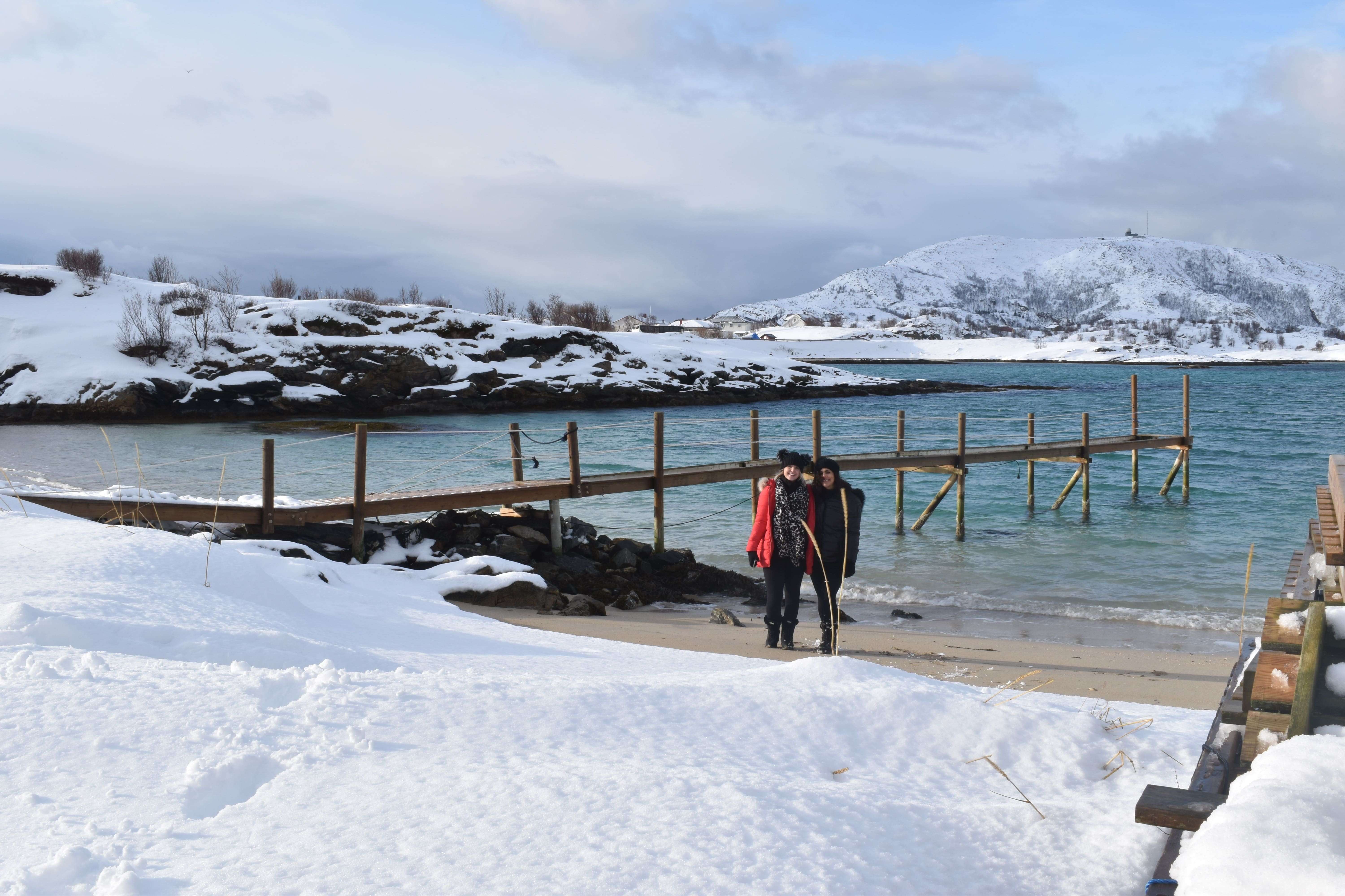 Two people stand on a snowy beach in winter clothing, with a wooden pier extending into the turquoise water. Snowy hills reminiscent of the Tromsø to Senja landscape are visible in the background under a cloudy sky.