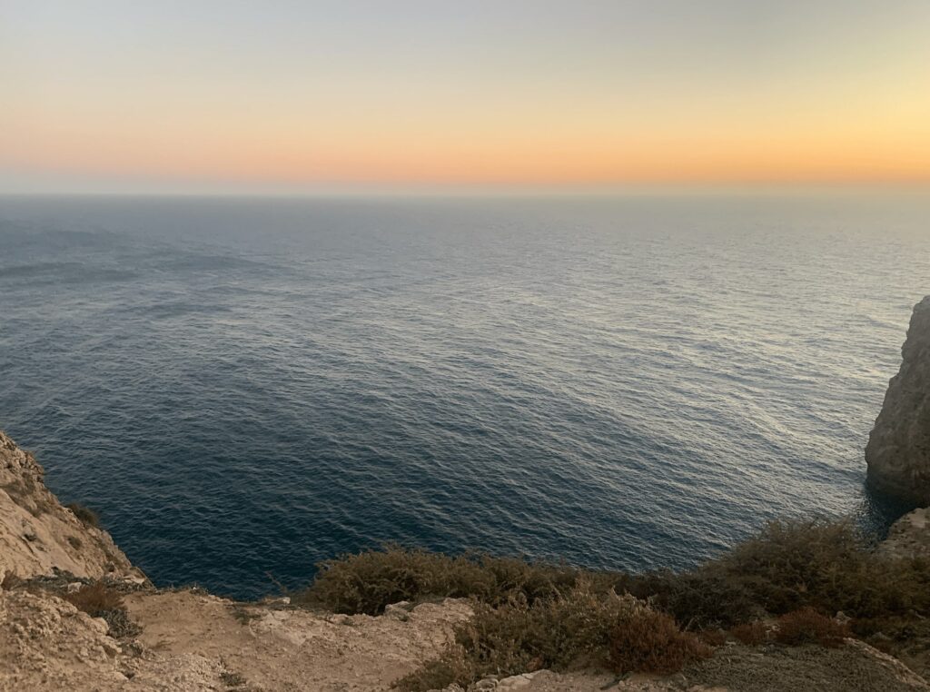 View from a rocky cliff overlooking the calm blue sea at sunset, with soft orange and yellow hues fading into the sky—one of the top things to do in Sagres. Sparse dry shrubs dot the tranquil foreground scenery.