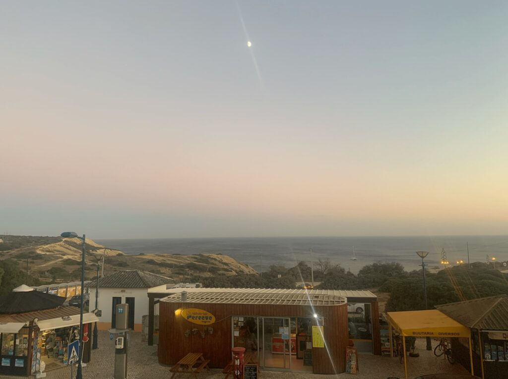 A coastal scene at dusk with small shops in the foreground, including a pizza place, and a calm sea beyond. The pastel sky and visible moon hint at the relaxing things to do in Sagres as evening falls.