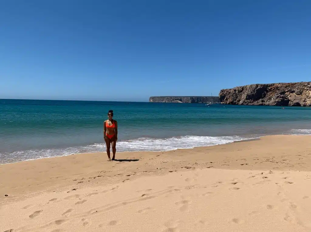 A person in a red swimsuit stands on a sandy beach near the shoreline in Sagres, with turquoise waves, rocky cliffs, and a clear blue sky—a perfect spot for those exploring things to do in Sagres.