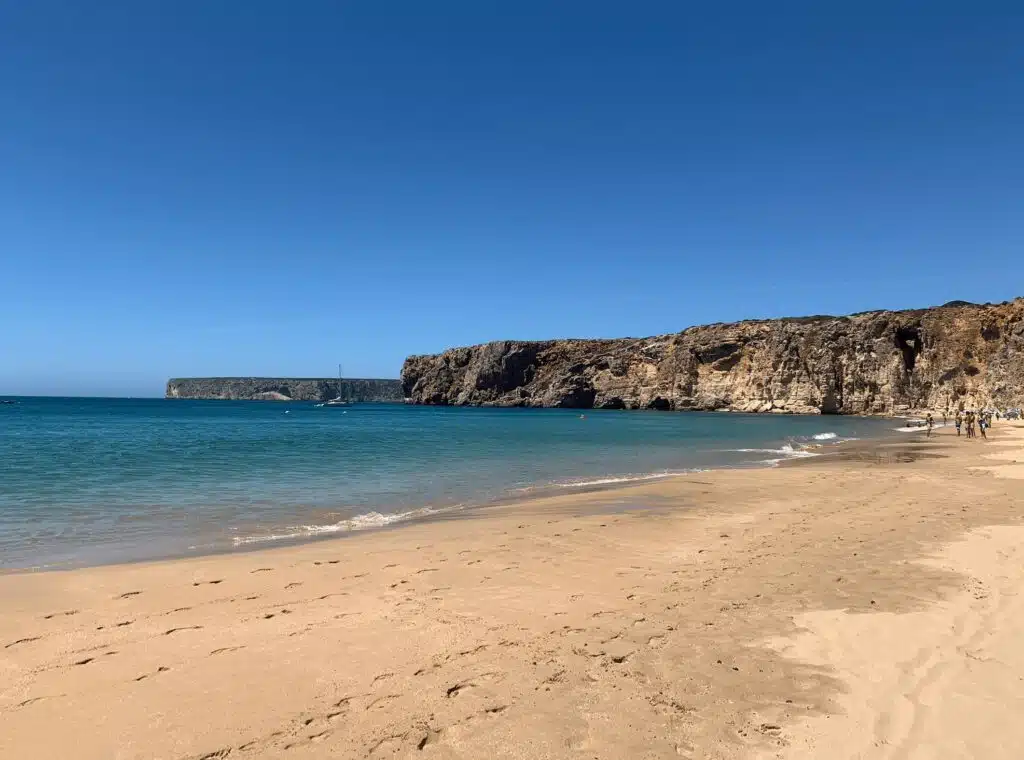 A sandy beach with clear blue water, rocky cliffs in the background, and a bright blue sky. A few people are visible near the water and along the shore, enjoying one of the top things to do in Sagres.