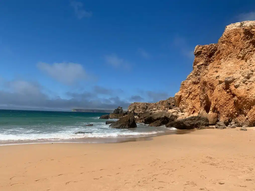A sandy beach with gentle waves, turquoise water, and large orange-brown rocky cliffs under a clear blue sky with scattered clouds—one of the must-see things to do in Sagres.