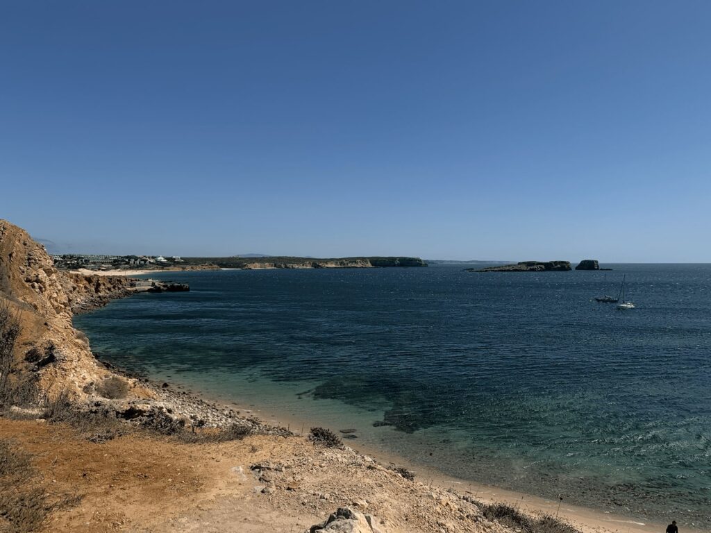 Rocky coastline with cliffs and dry grass in the foreground, clear blue ocean water, a distant sailboat, and a small group of islands under a clear sky—perfect for discovering things to do in Sagres.