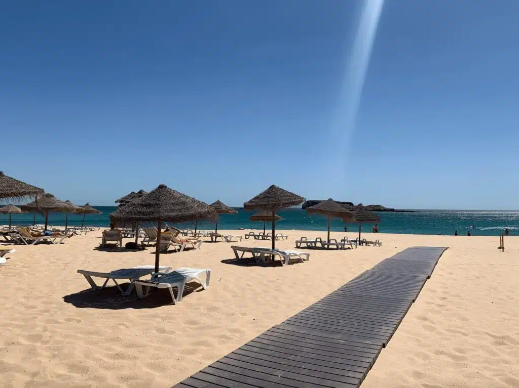 A sunny beach with straw parasols and empty lounge chairs on the sand. A wooden walkway leads toward the turquoise sea under a clear blue sky—one of the top things to do in Sagres for relaxation and stunning views.