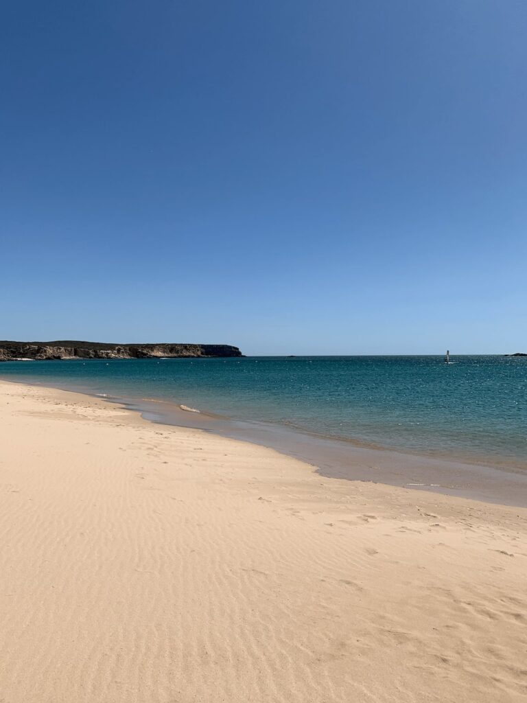A sandy beach with gentle waves and clear turquoise water under a bright blue sky—one of many tranquil spots among the things to do in Sagres. In the distance, low rocky cliffs border the coastline. The beach appears calm and empty.